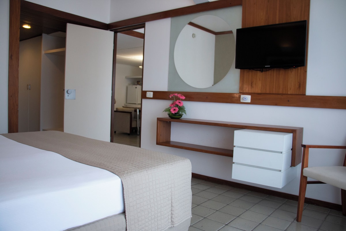 A hotel room with a bed, a TV mounted on the wall, a mirror, a wooden desk with drawers, and a small shelf with flowers.