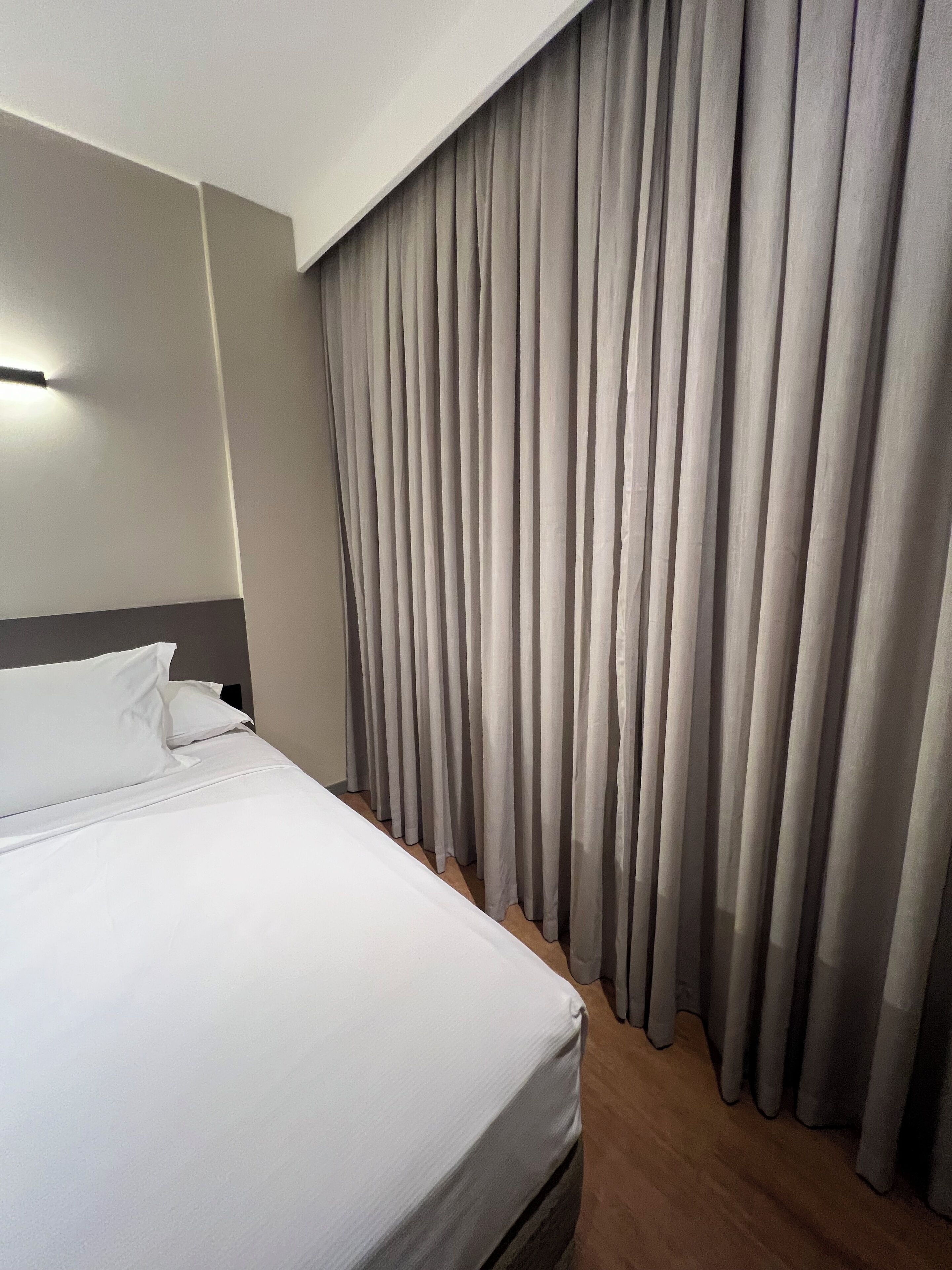 A hotel room with a neatly made bed, a headboard, and a large window with curtains.