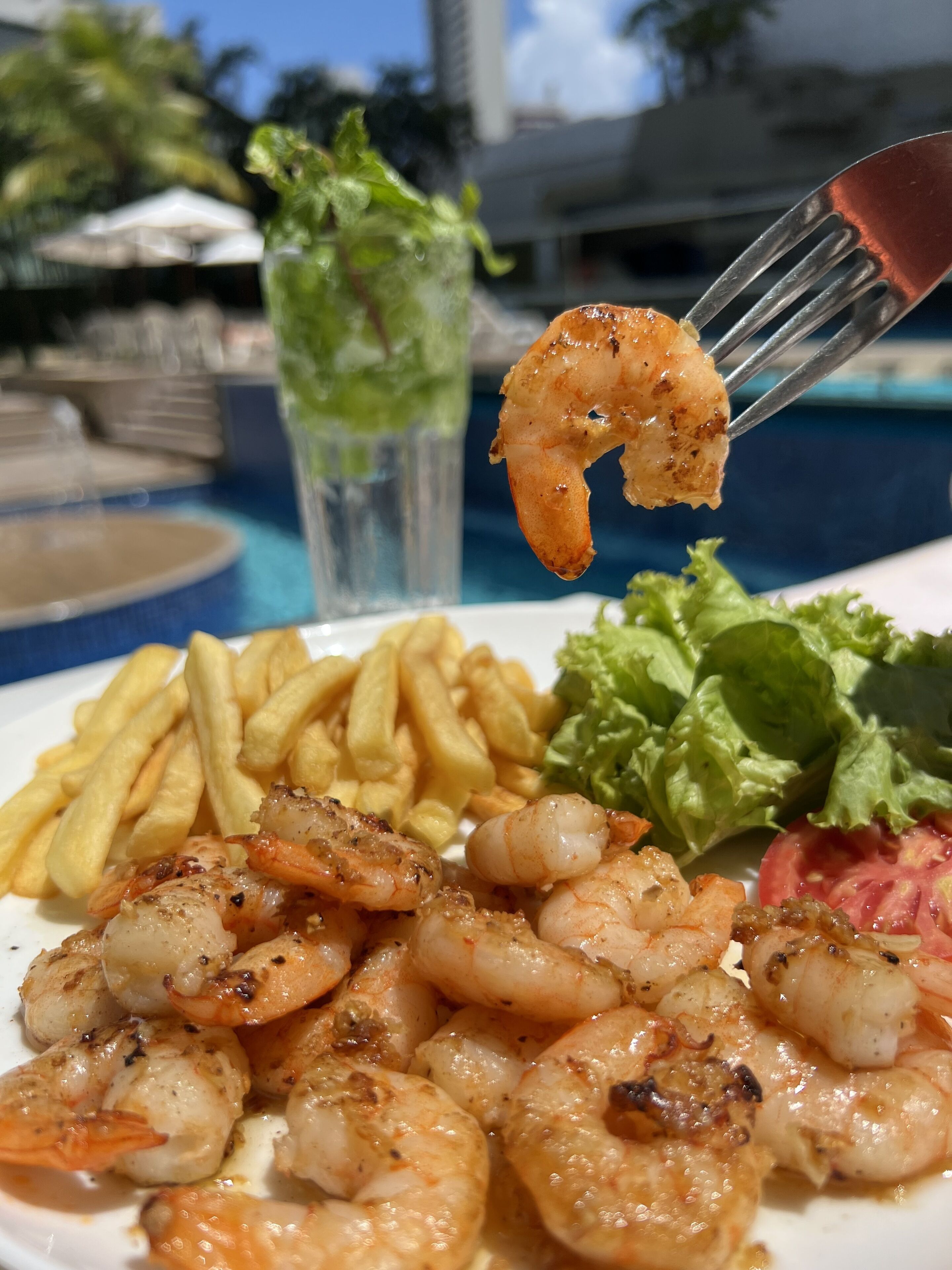 Un plato de camarones a la parrilla, papas fritas y una ensalada con un tenedor sosteniendo un camarón.