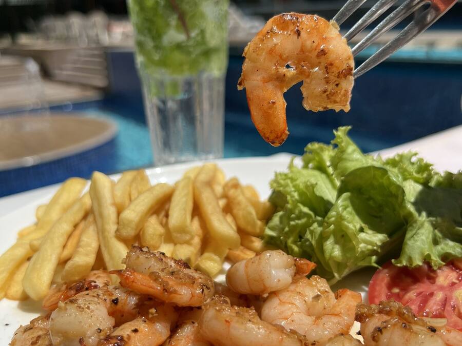 Un plato de camarones a la parrilla, papas fritas y una ensalada con un tenedor sosteniendo un camarón.