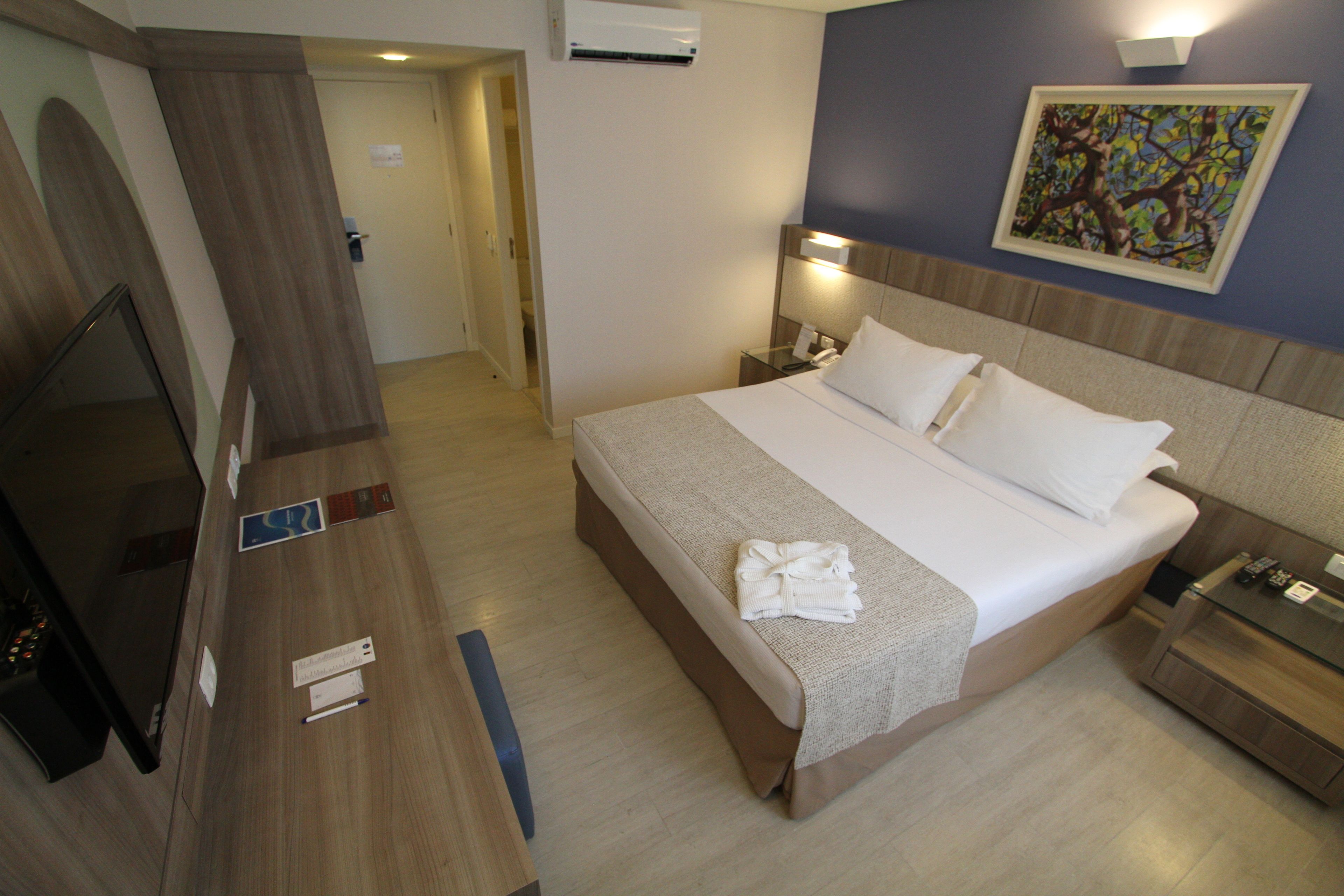 A hotel room with a bed, bedside table, TV, and a painting on the wall.