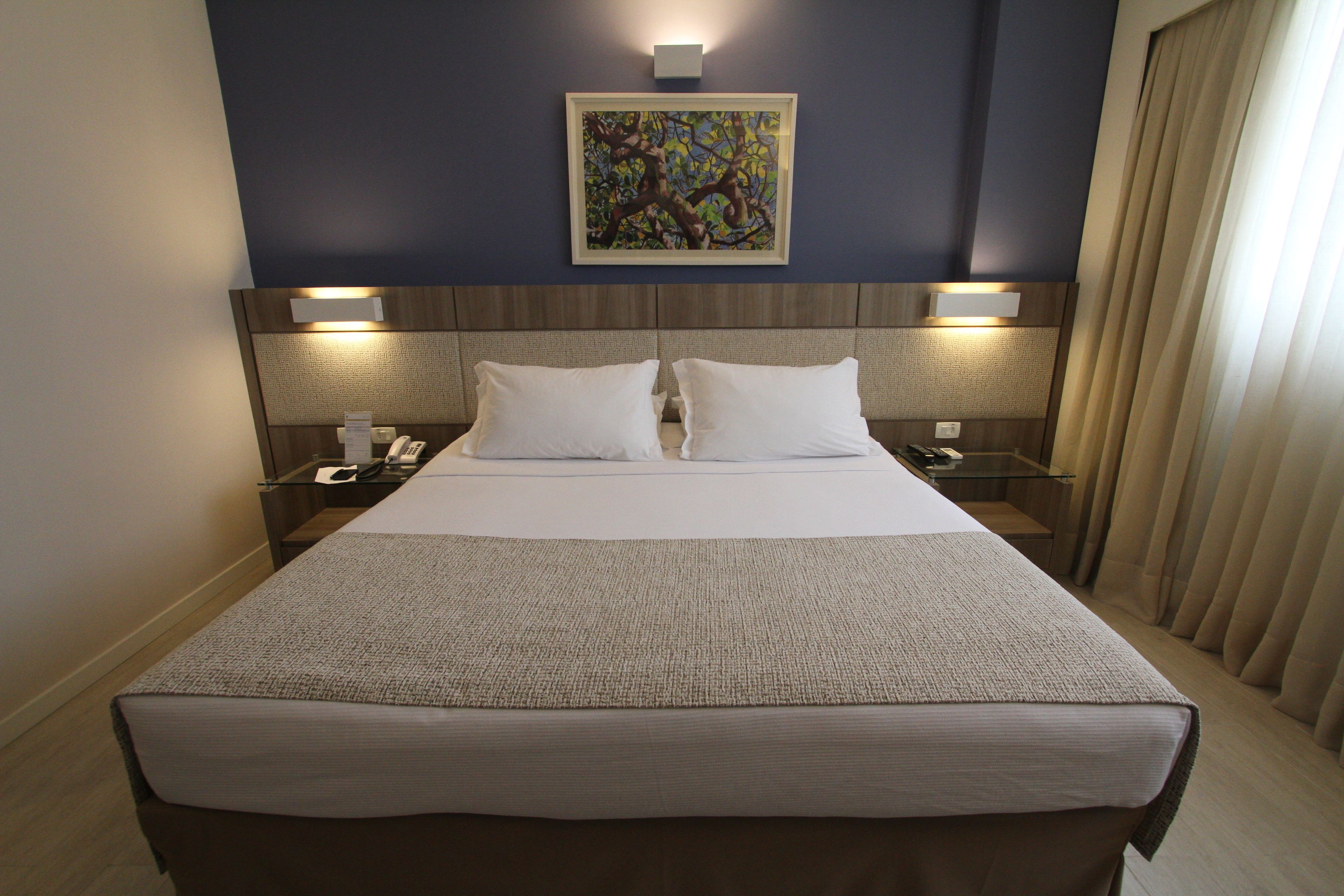 A hotel room with a large bed, two bedside tables, a nightstand, a painting on the wall, and a window with curtains.