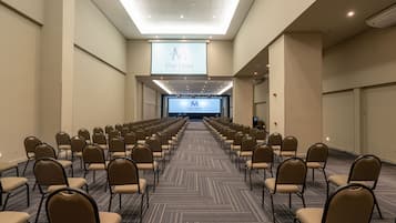 Une salle de conférence avec des rangées de chaises, un grand écran à l’avant et un sol recouvert de moquette.