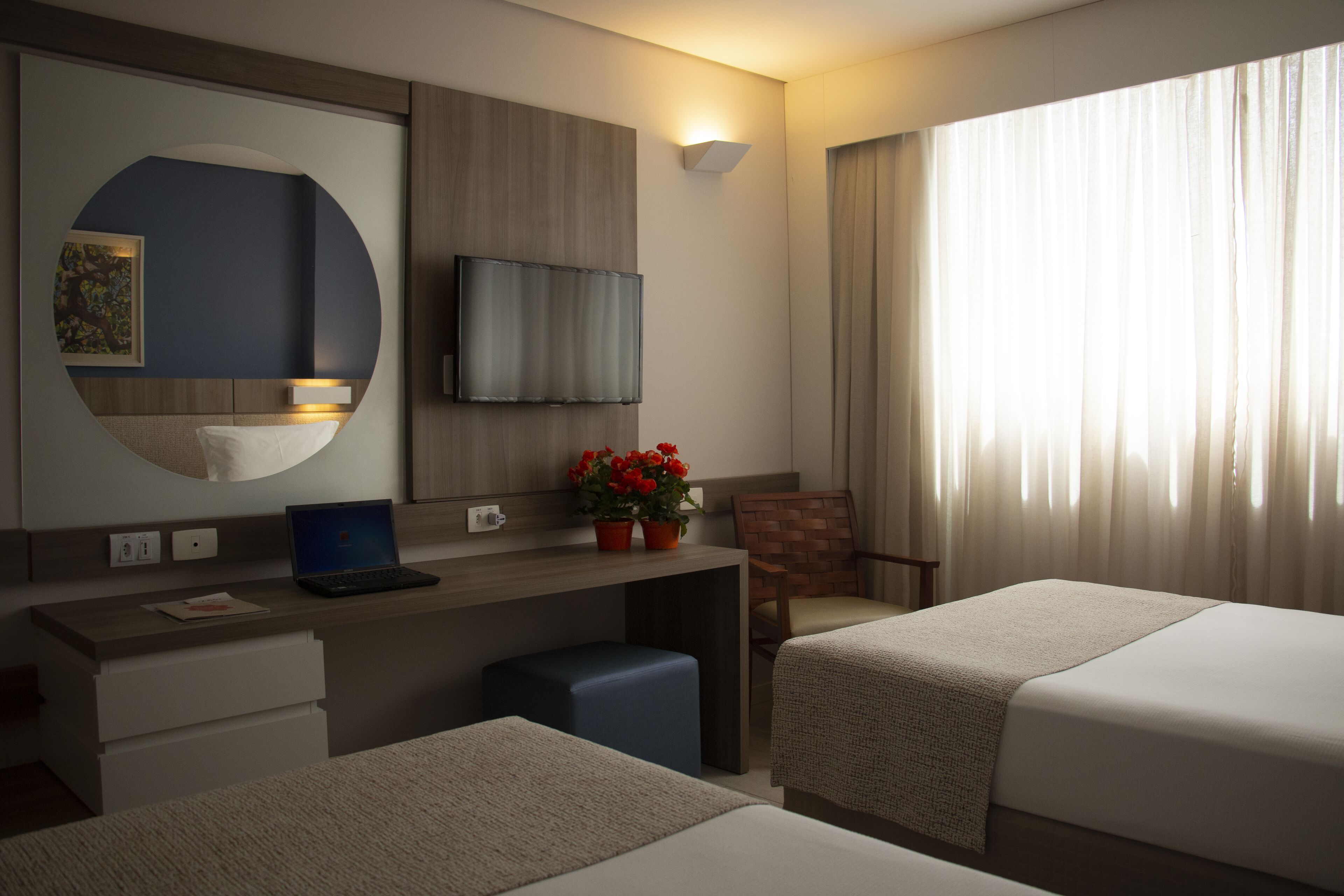 Habitación de hotel con dos camas, un escritorio con una laptop, un televisor montado en la pared, una planta en maceta y una ventana con cortinas.