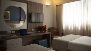 Habitación de hotel con dos camas, un escritorio con una laptop, un televisor montado en la pared, una planta en maceta y una ventana con cortinas.