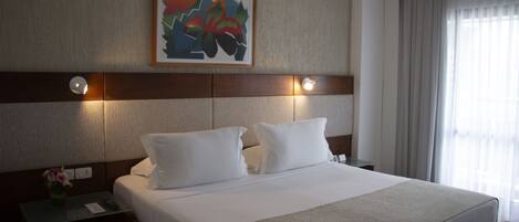 Quarto de hotel com uma cama grande, mesa de cabeceira com um vaso de flores e um quadro na parede.