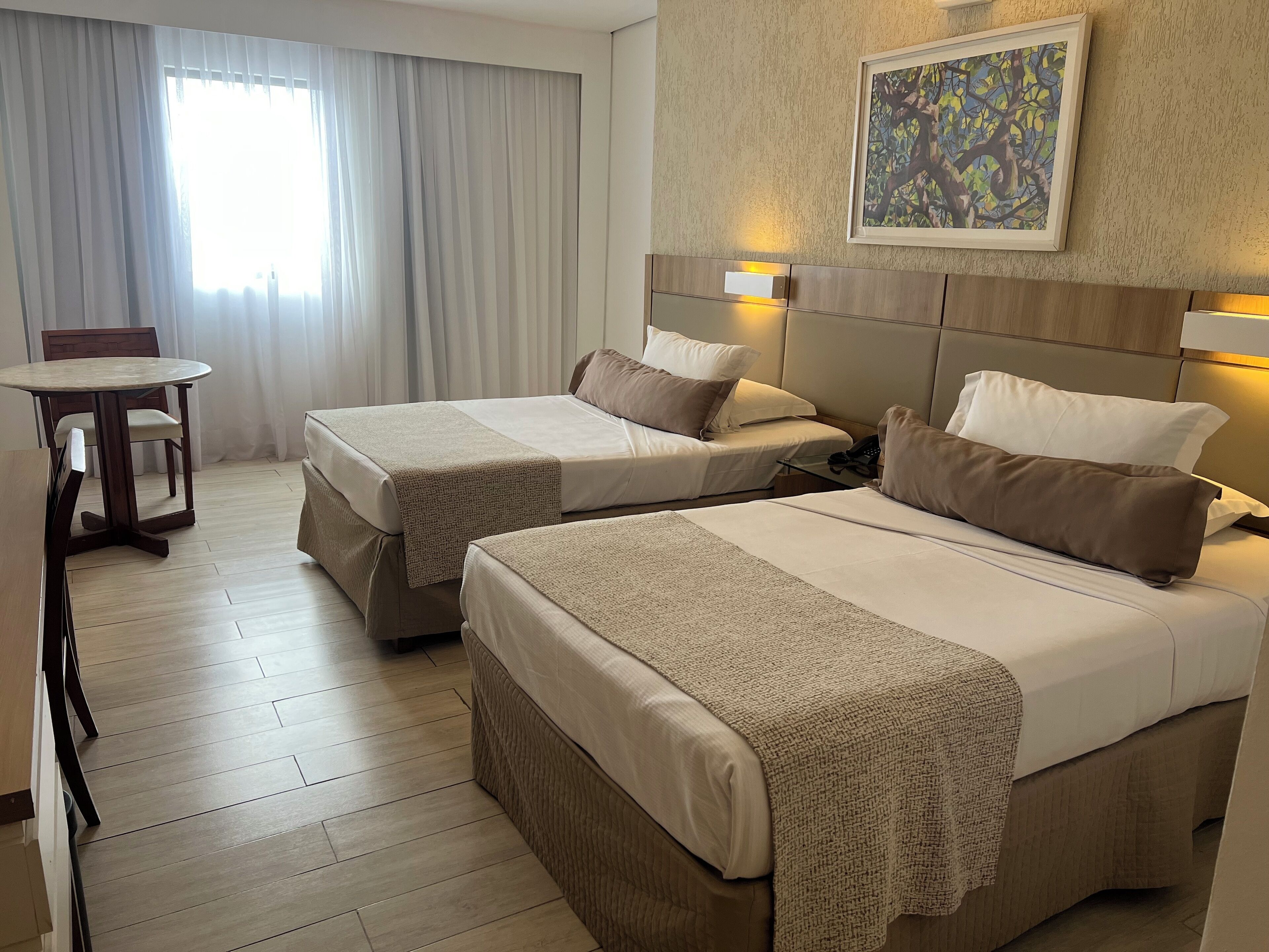 A hotel room with two beds, a dining table, and a painting on the wall.