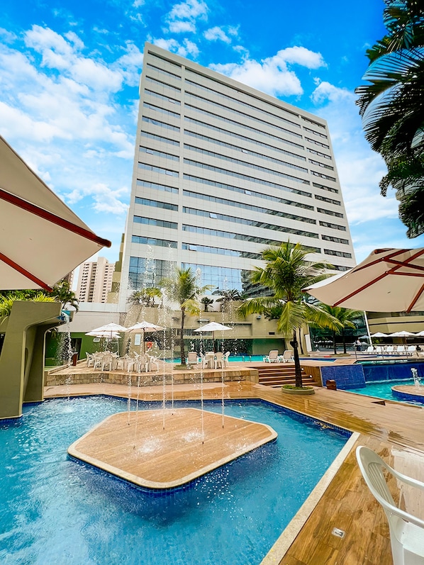 Una piscina con una fuente de agua, rodeada de sillas de descanso y sombrillas, con un edificio alto al fondo.