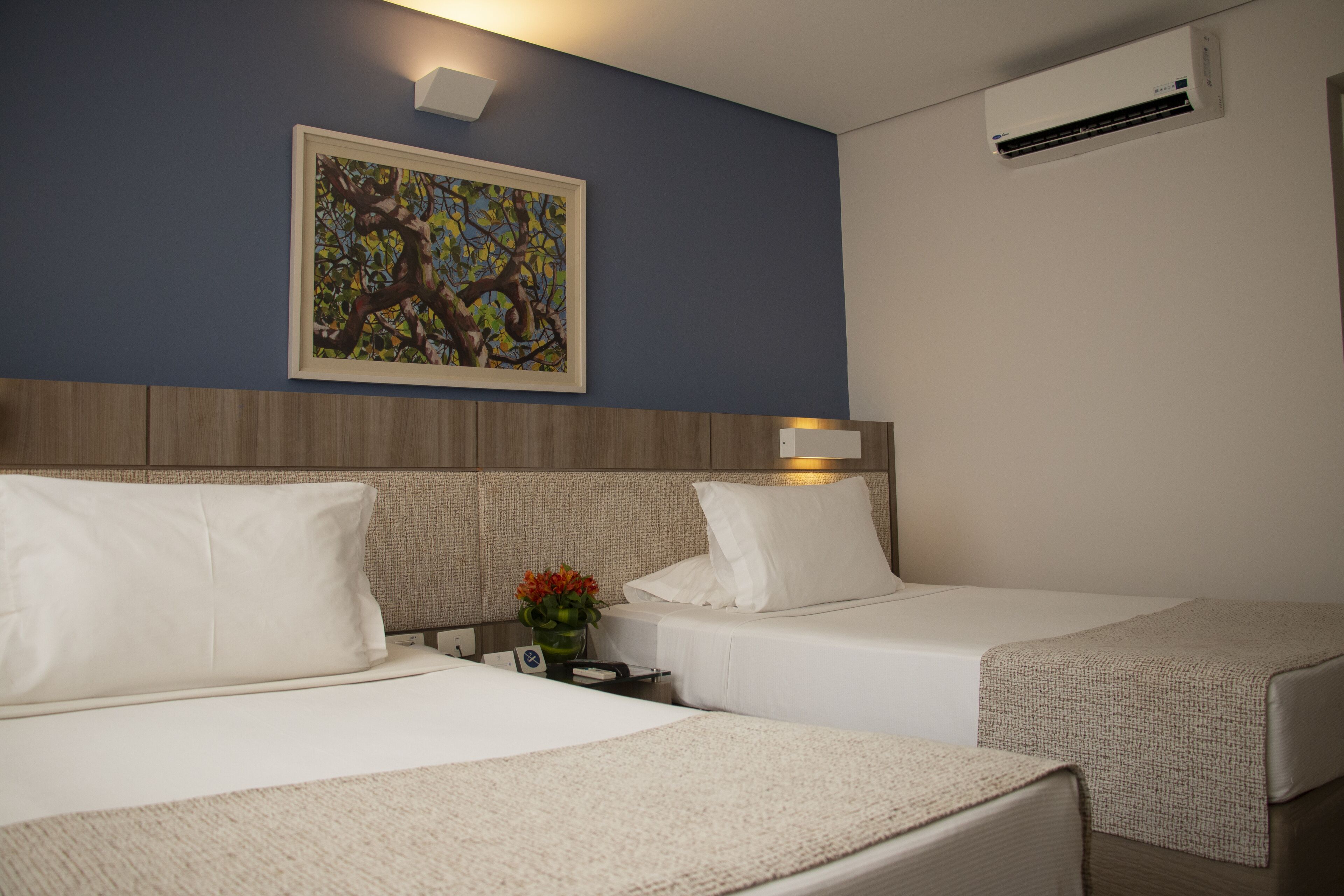 A hotel room with two beds, a painting on the wall, an air conditioner, and a nightstand with a vase of flowers.