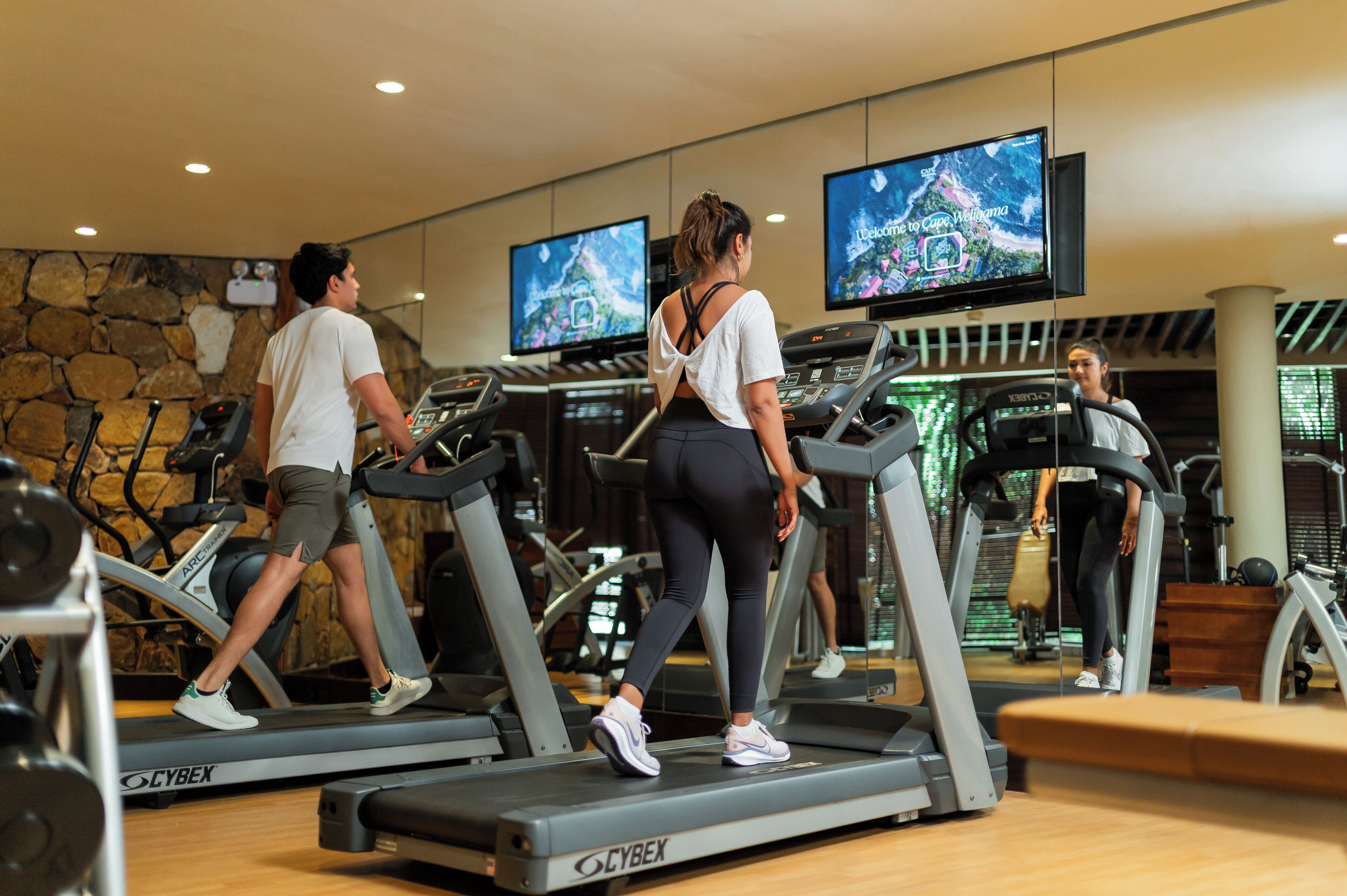 Une salle de sport équipée de tapis de course, dotée d’un mur en pierre et de téléviseurs diffusant une carte.