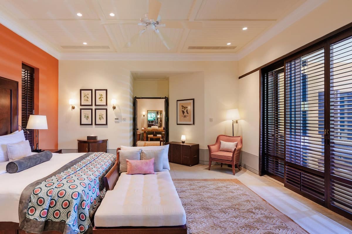 A hotel room with a large bed, a sofa, a desk, and a chair. There is a ceiling fan, a window with blinds, and framed pictures on the wall.