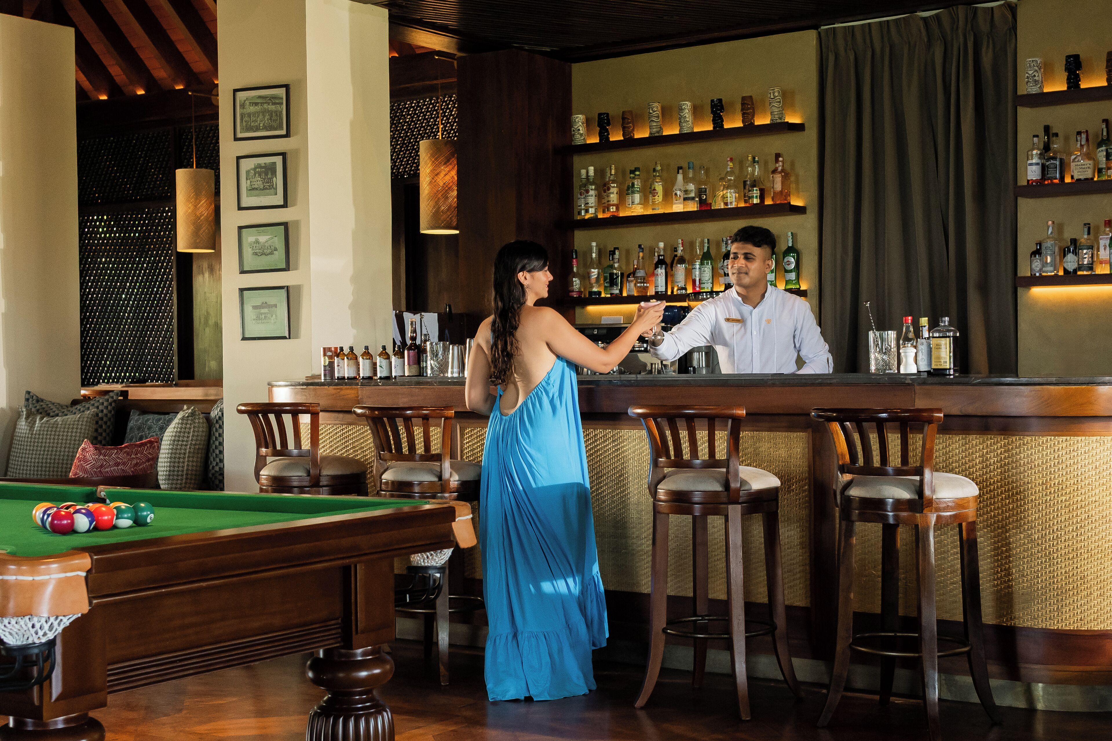 Een bar met een pooltafel, barstoelen en een barman die een klant bedient.