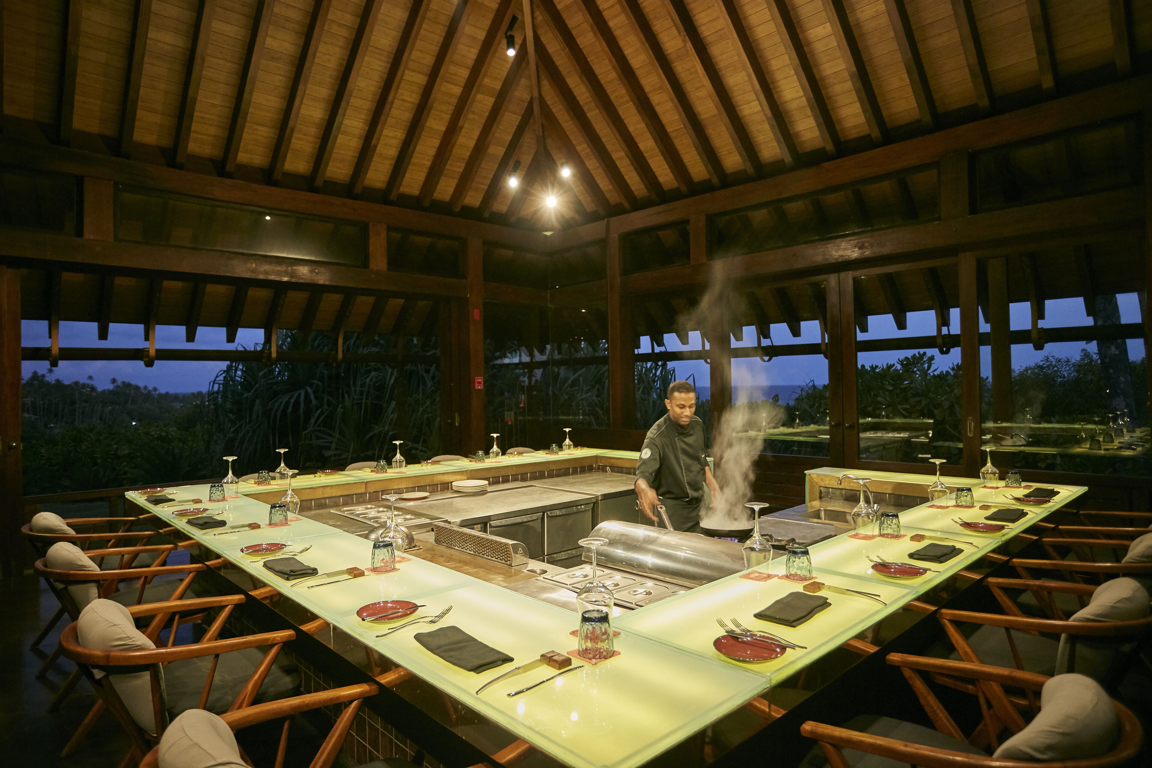 A long dining table set for a meal with a chef preparing food in the center.