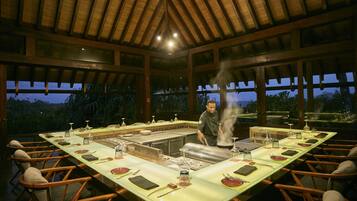 A long dining table set for a meal with a chef preparing food in the center.