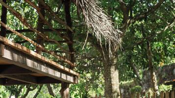 Two individuals sitting on a wooden deck outdoors, surrounded by lush greenery and a wooden structure overhead.