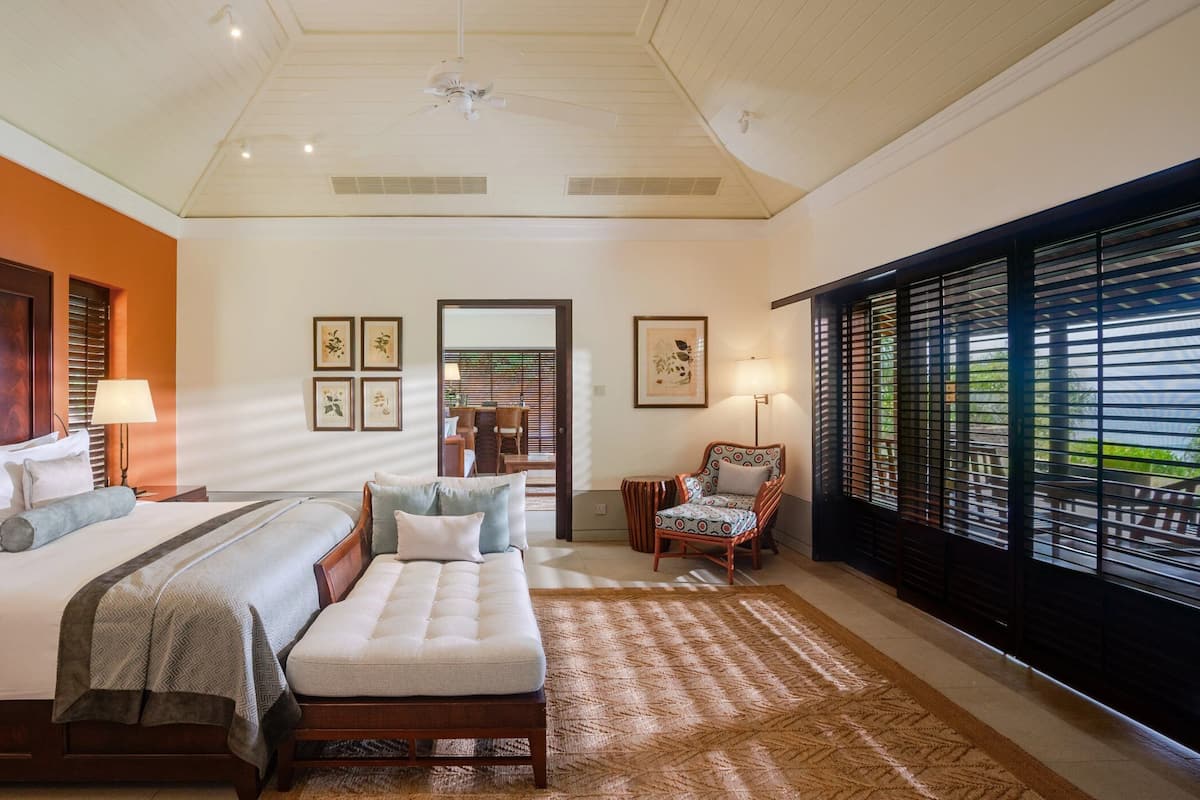 A hotel room with a large bed, a sofa, a chair, and a window with blinds. There are framed pictures on the wall and a ceiling fan.