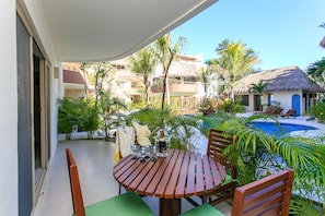 A wooden table with chairs on a balcony overlooking a swimming pool and thatched-roof buildings.