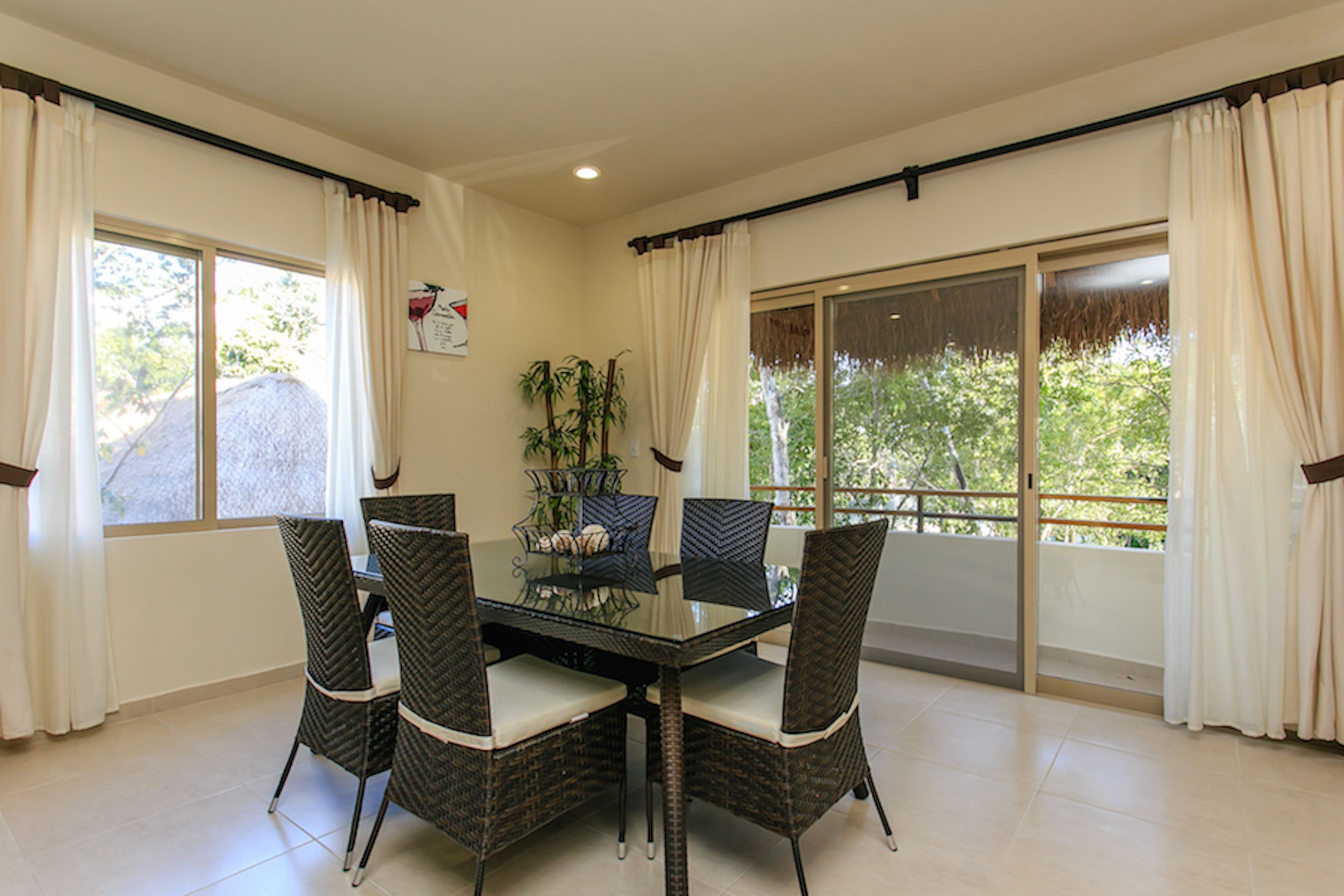 A dining area with a glass table surrounded by wicker chairs, large windows with curtains, and a view of greenery outside.