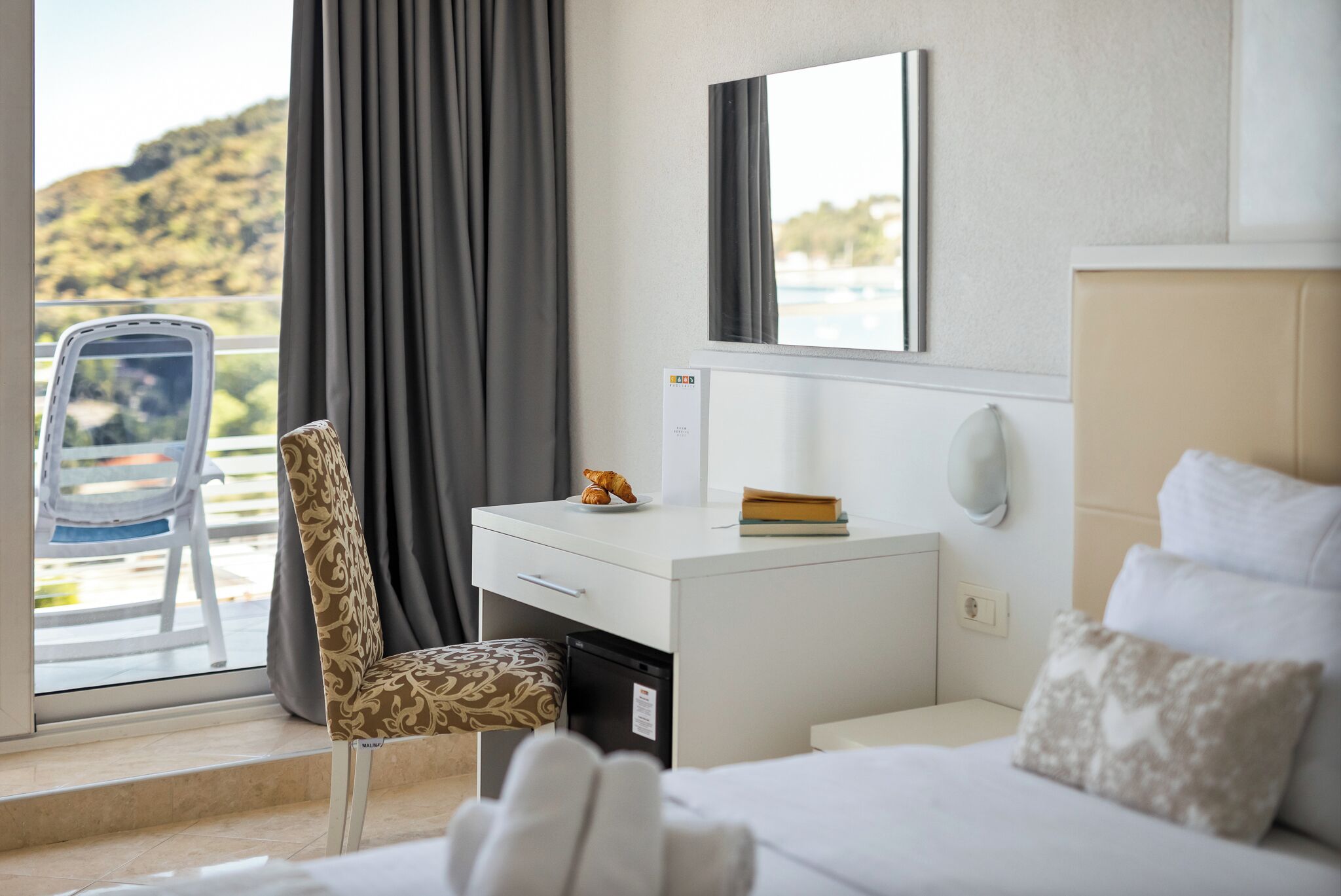 A hotel room with a bed, a desk with a computer, a chair, a window with curtains, and a view of the outdoors.