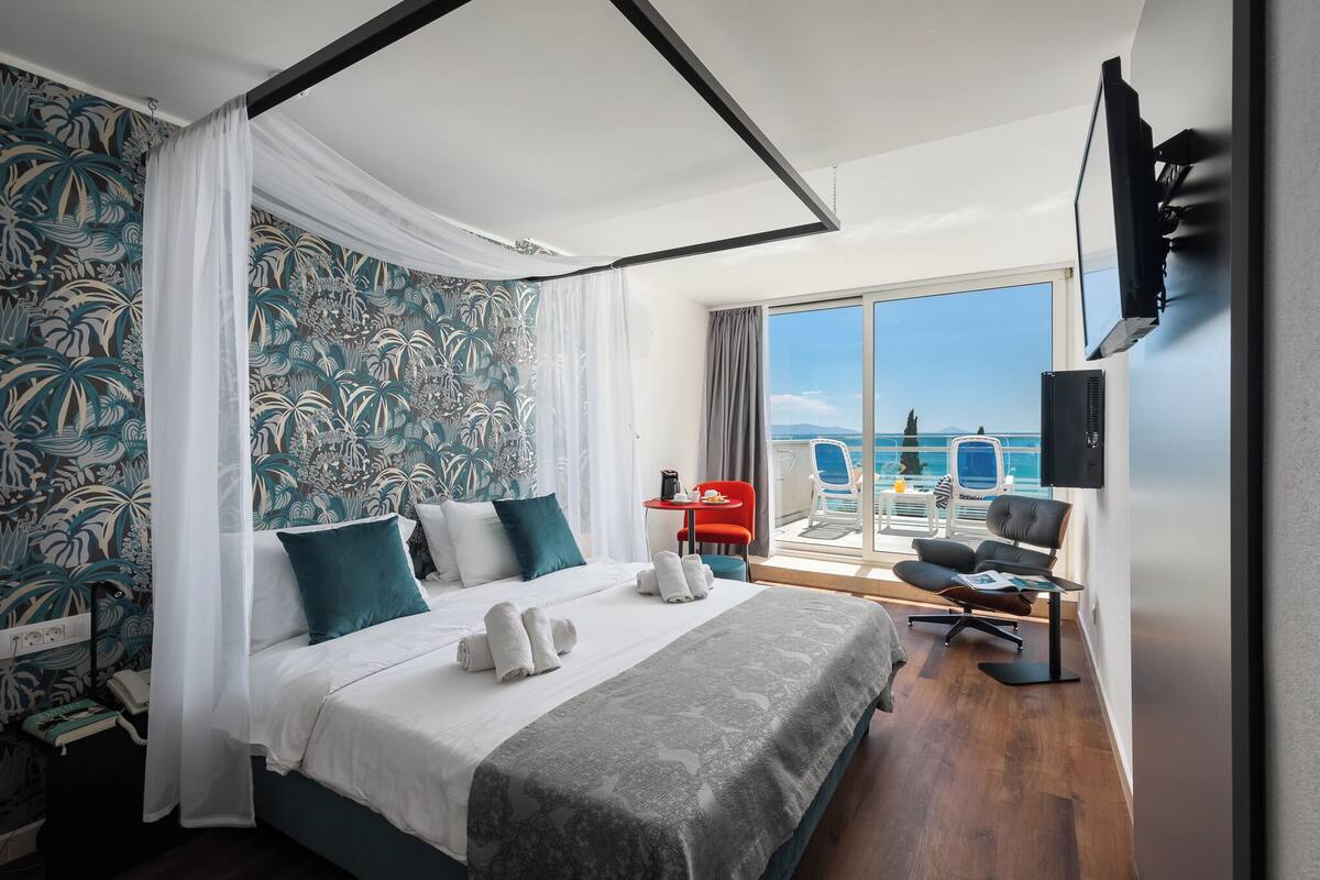 A hotel room with a bed, a balcony with a view of the sea, a TV mounted on the wall, and a small table with a chair.