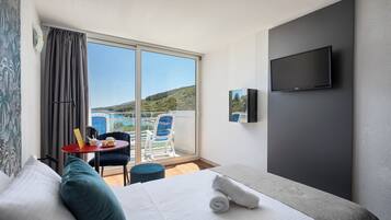 A hotel room with a bed, a TV, a balcony with a chair, and a table with a cup and a book.