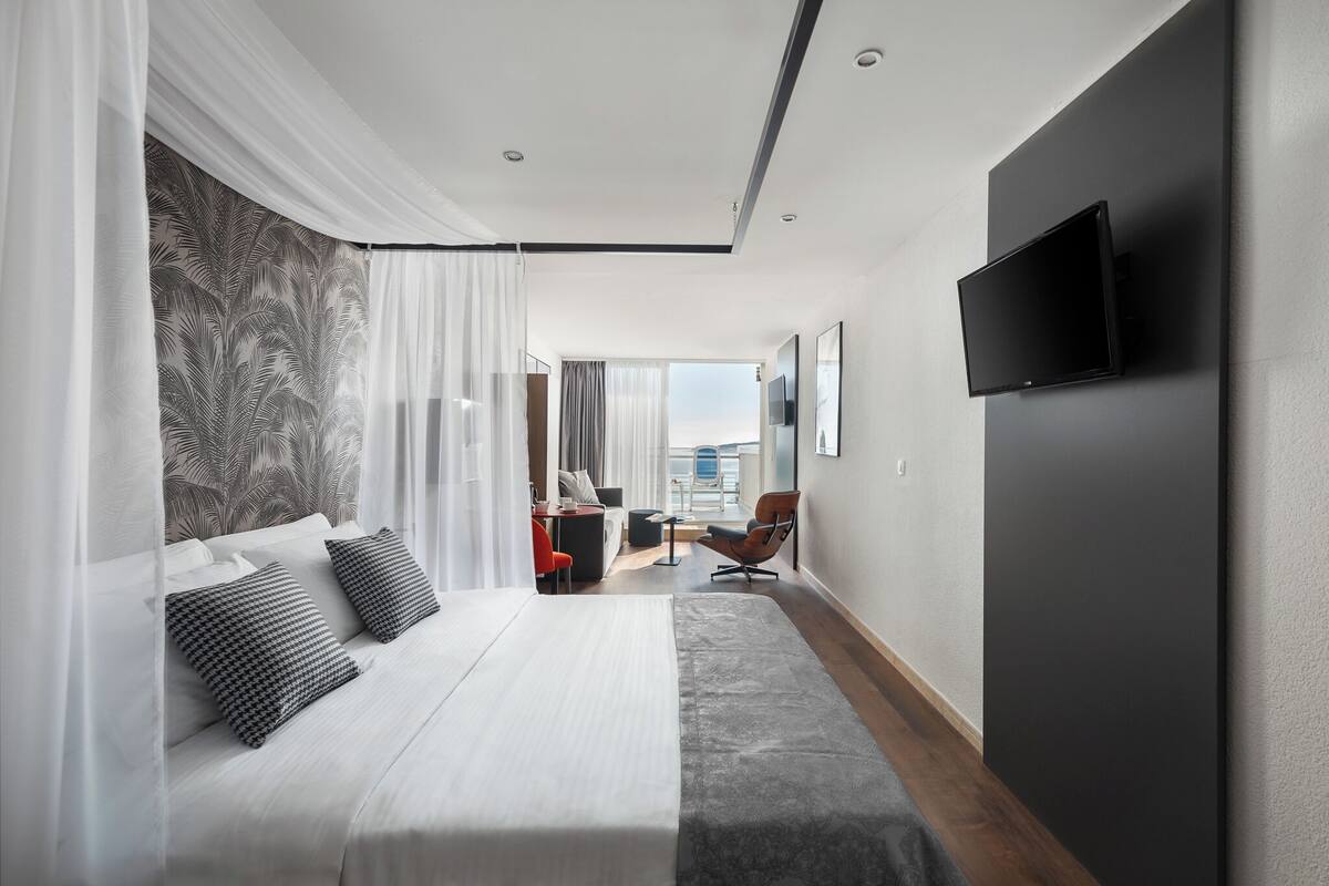 A hotel room with a bed, a desk, a chair, a TV, and a view of the sea.