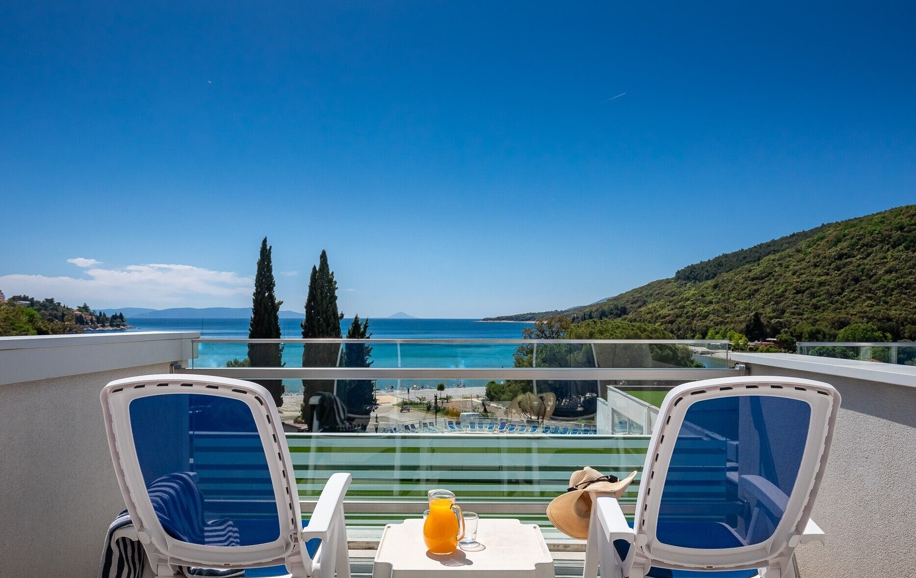 Ein Balkon mit zwei Stühlen, einem Tisch und Blick auf Meer und Berge.