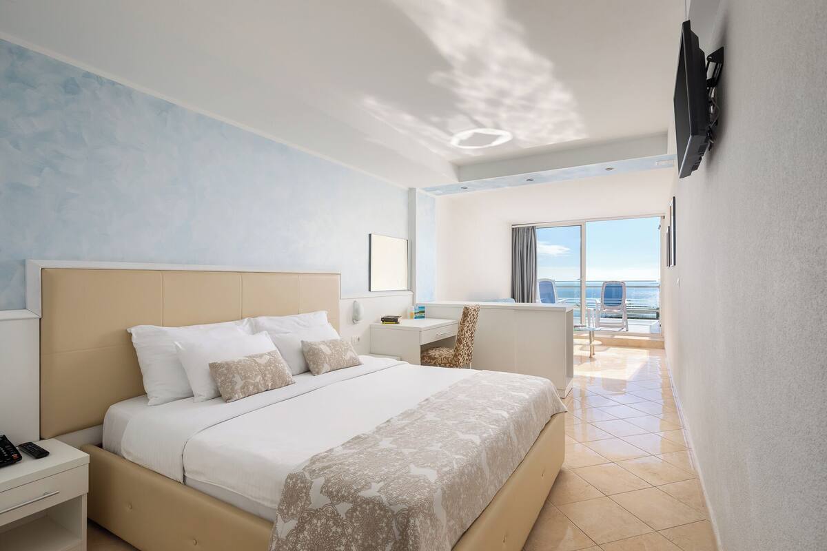 A modern hotel room with a large bed, a desk, and a view of the sea.