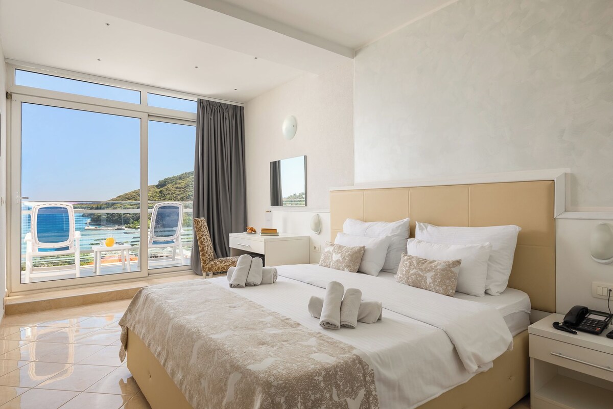 A hotel room with a large bed, a bedside table with a phone, and a view of a pool and mountains through large windows.