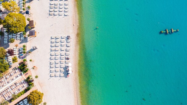 An aerial view of a beach with rows of sun loungers and umbrellas, a clear turquoise sea, and a small boat in the water. - MASLINICA Mimosa Lido Palace Hotel (Labin)