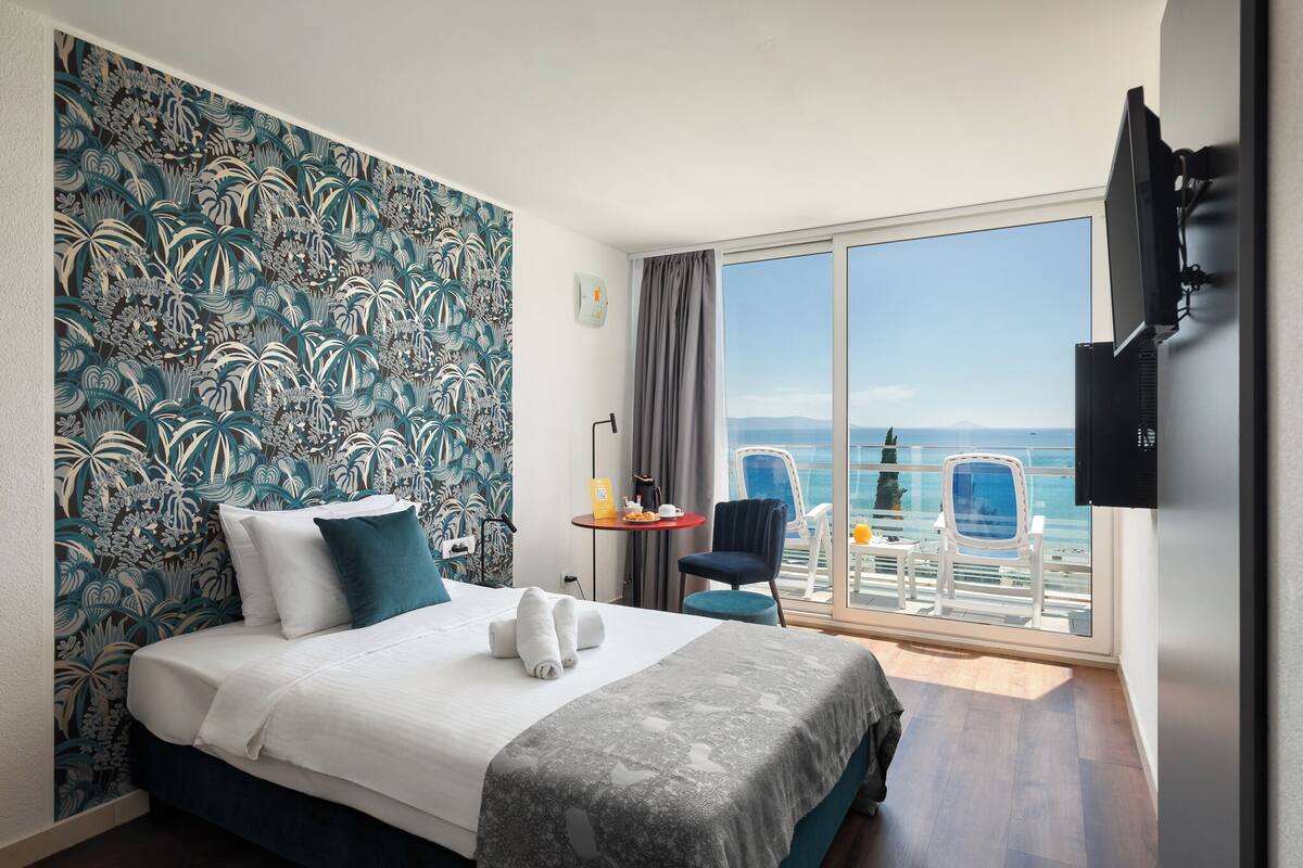 A hotel room with a bed, a desk, a chair, a TV, and a balcony with a view.