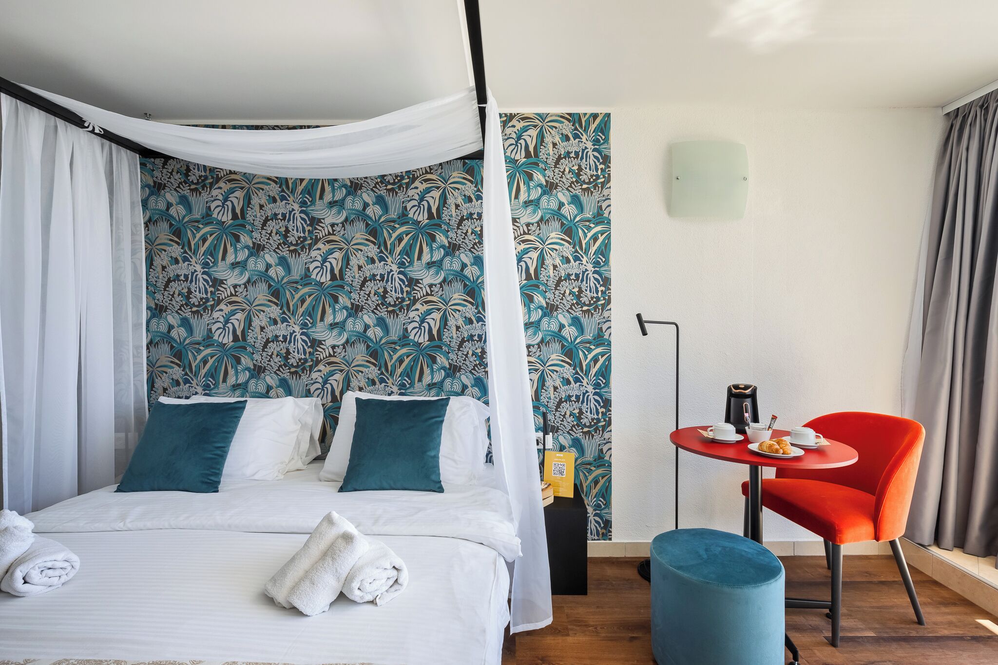 A bed with white linens, teal pillows, and a canopy, a small round table with a red chair, a blue ottoman, and a patterned wall.