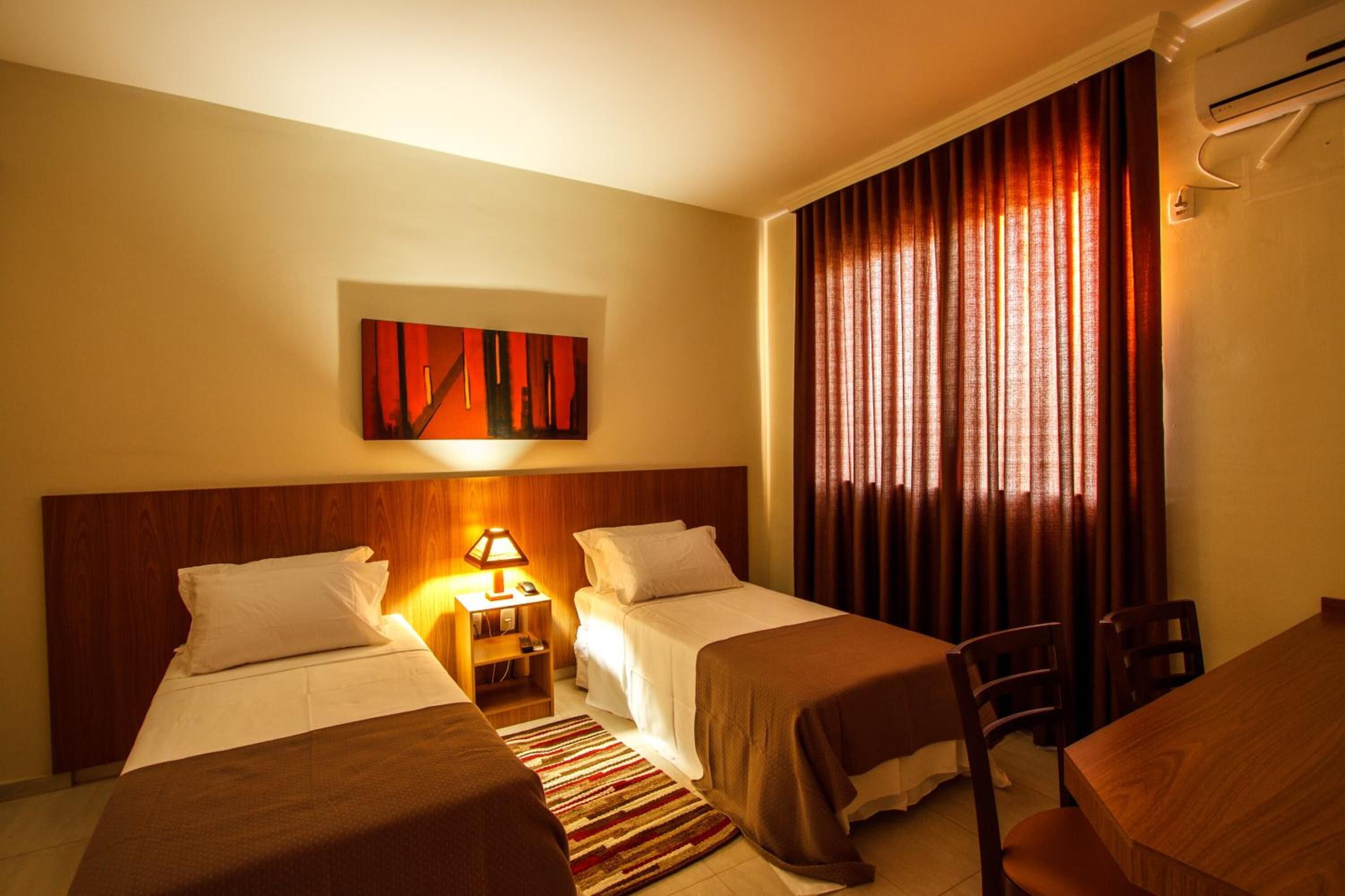 A hotel room with two beds, a wooden headboard, a nightstand with a lamp, a painting on the wall, and a large window with curtains.