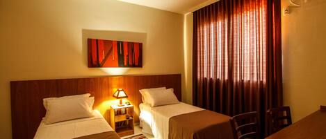 A hotel room with two beds, a wooden headboard, a nightstand with a lamp, a painting on the wall, and a large window with curtains.