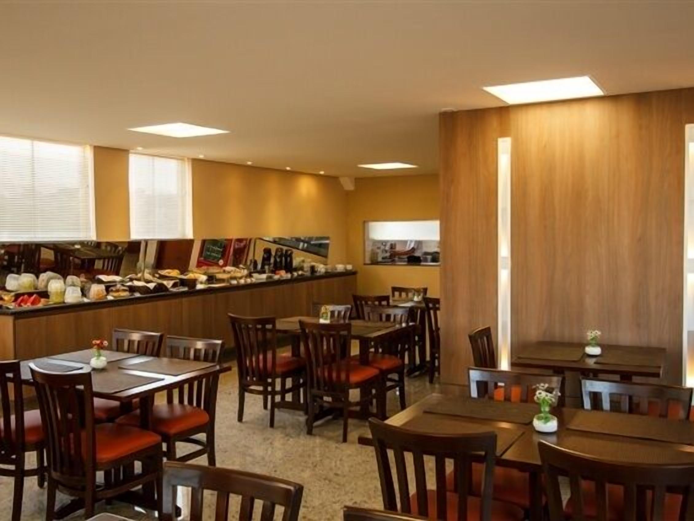 Un comedor de hotel con mesas y sillas de madera, una barra con diversos alimentos y una cocina visible al fondo.