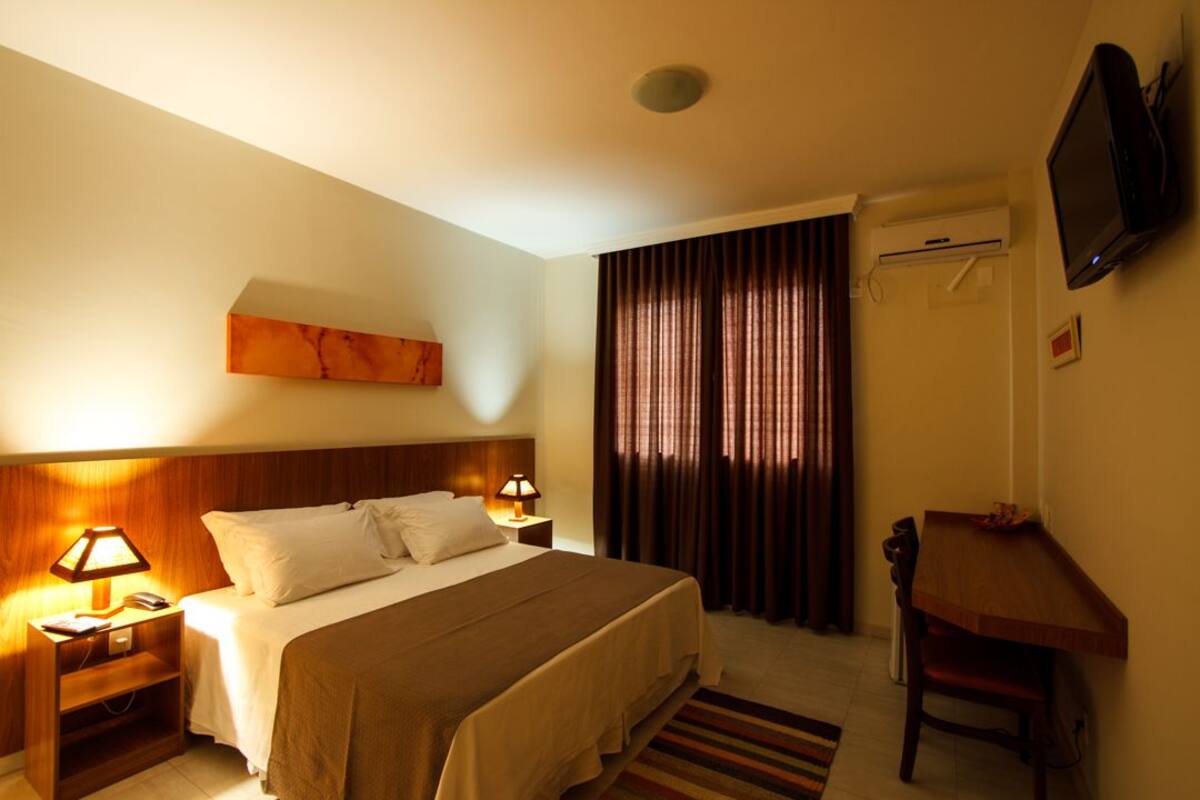 Una habitación de hotel con una cama, lámparas de noche, un escritorio, una silla, un televisor y una ventana con cortinas.