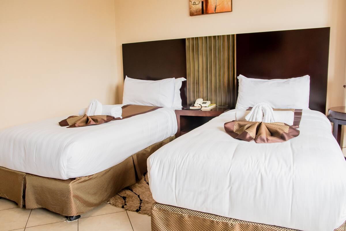 A hotel room with two beds, each with white linens and brown decorative pillows. There is a nightstand with a telephone and a lamp between the beds.