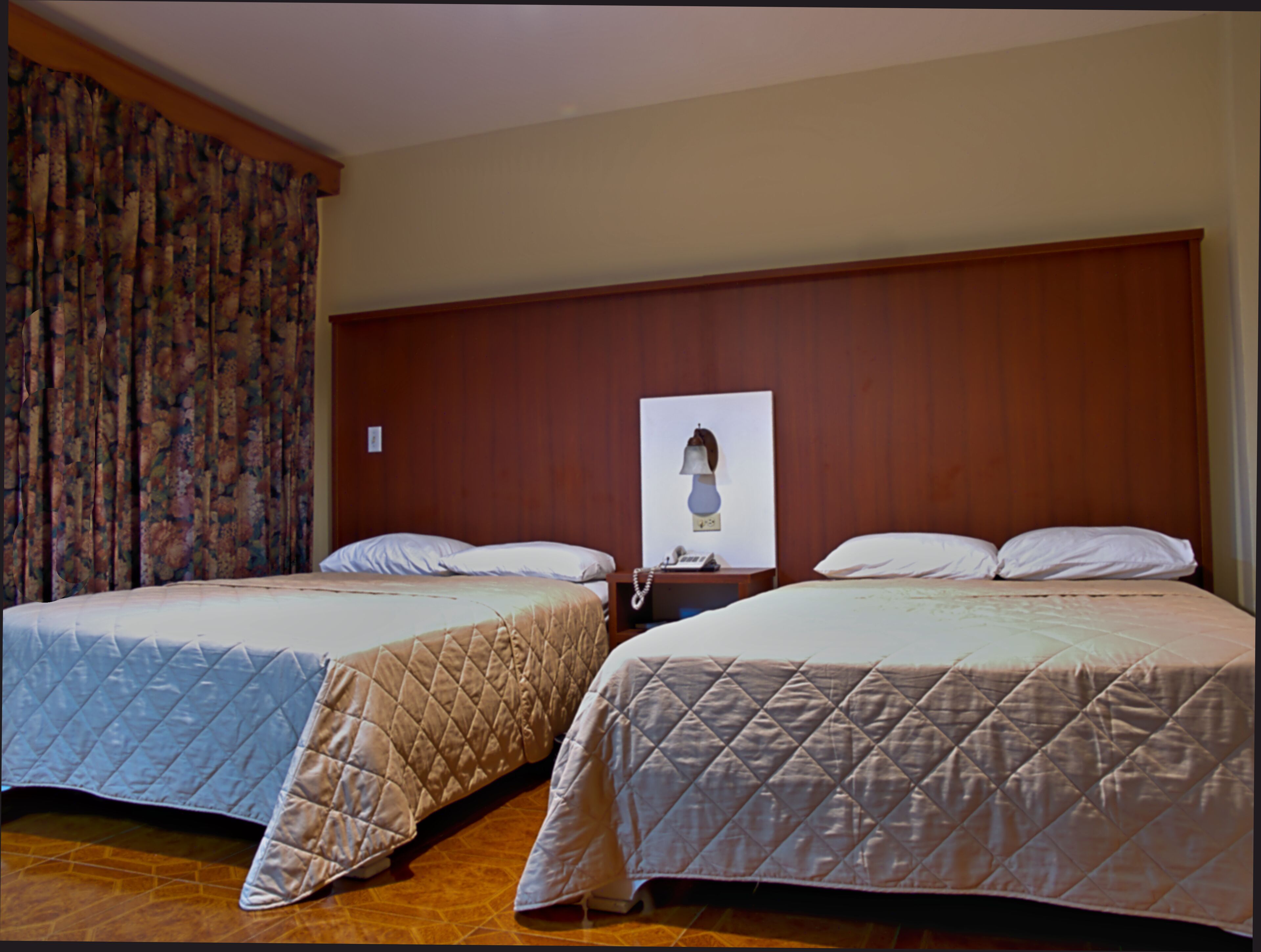 Dos camas matrimoniales con cabeceras de madera, una mesita de noche con teléfono y una cortina estampada con motivos florales.