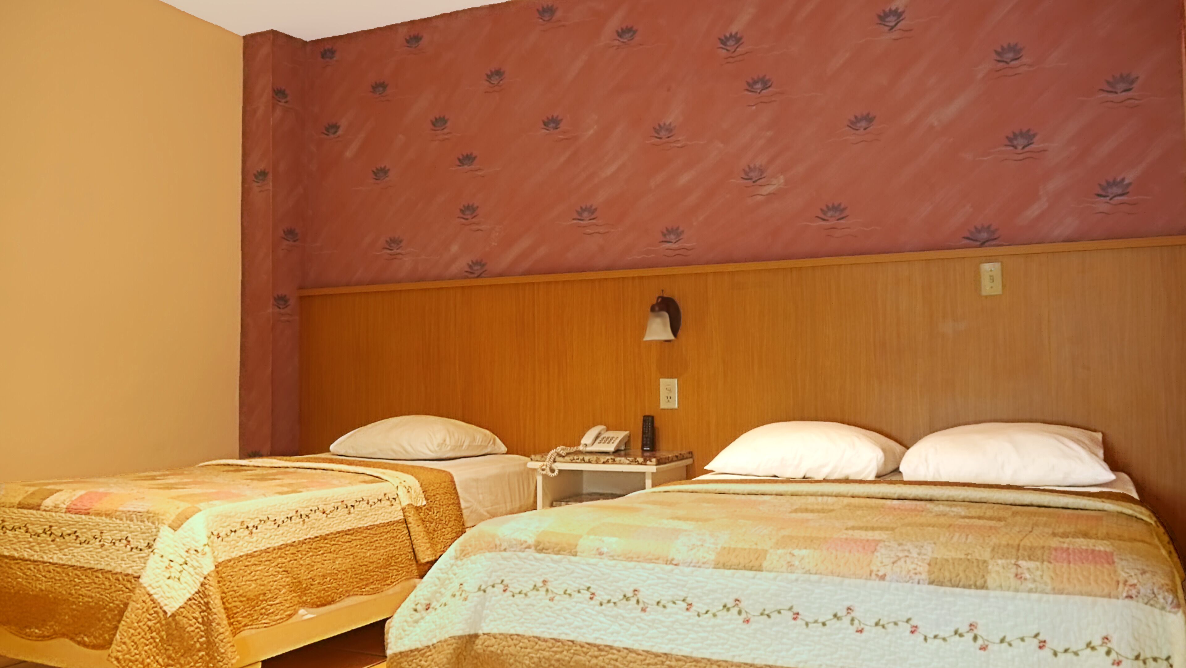 Une chambre d’hôtel avec deux lits, chacun doté d’une tête de lit en bois et d’un mur décoré d’un motif floral au-dessus. On y trouve une table de chevet avec une lampe et un téléphone.
