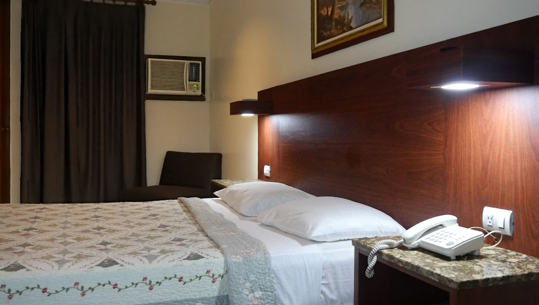 Habitación de hotel con una cama, teléfono en la mesita de noche, aire acondicionado de pared y un cuadro sobre la cama.
