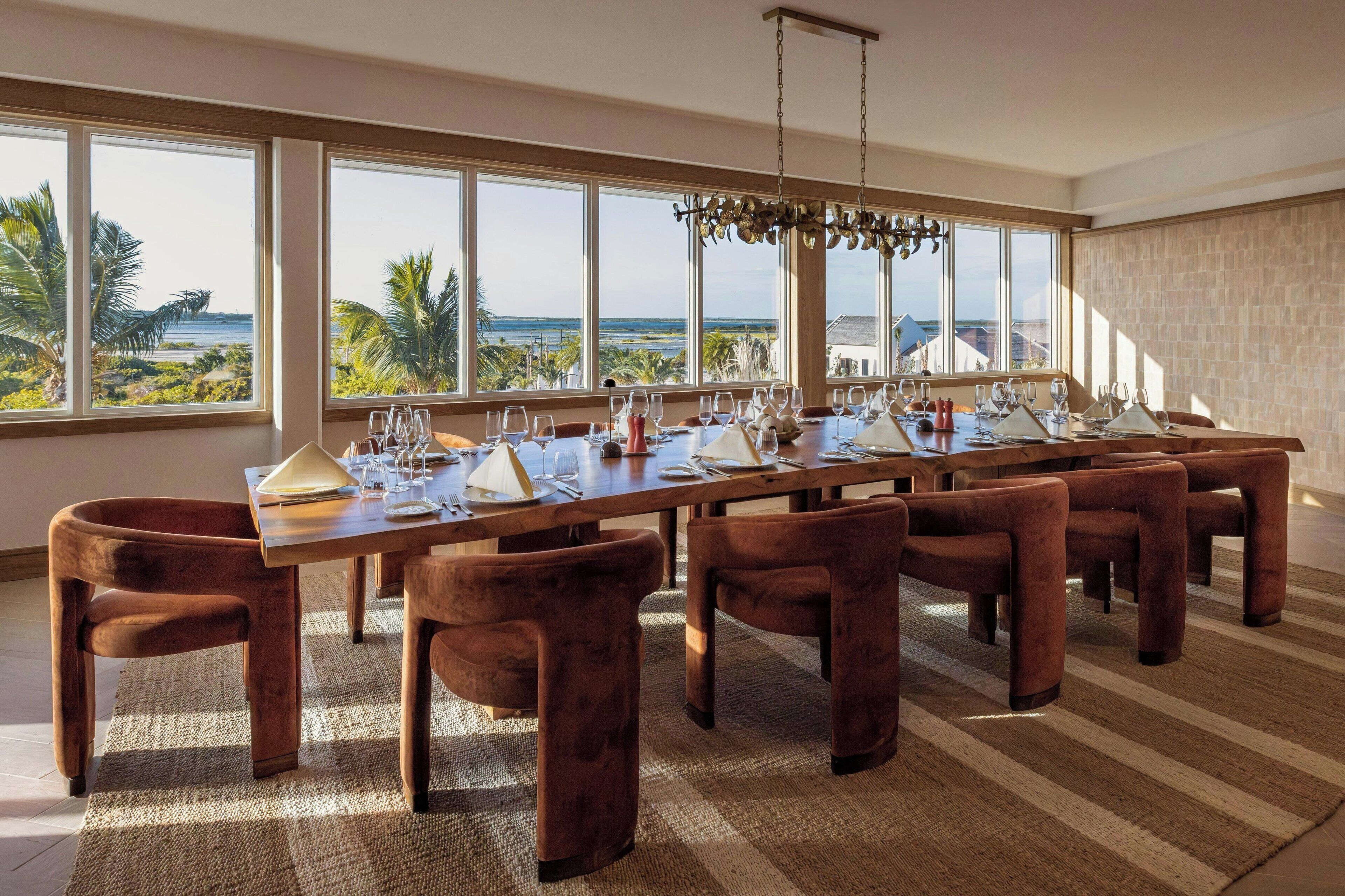 A dining room with a long table set for a meal, large windows with a view of the ocean, and a chandelier.