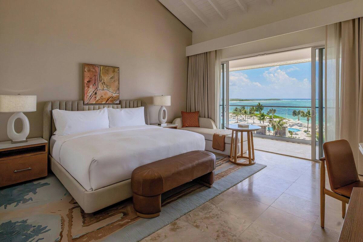 A hotel room with a large bed, a bench, a desk, and a view of the beach and ocean.