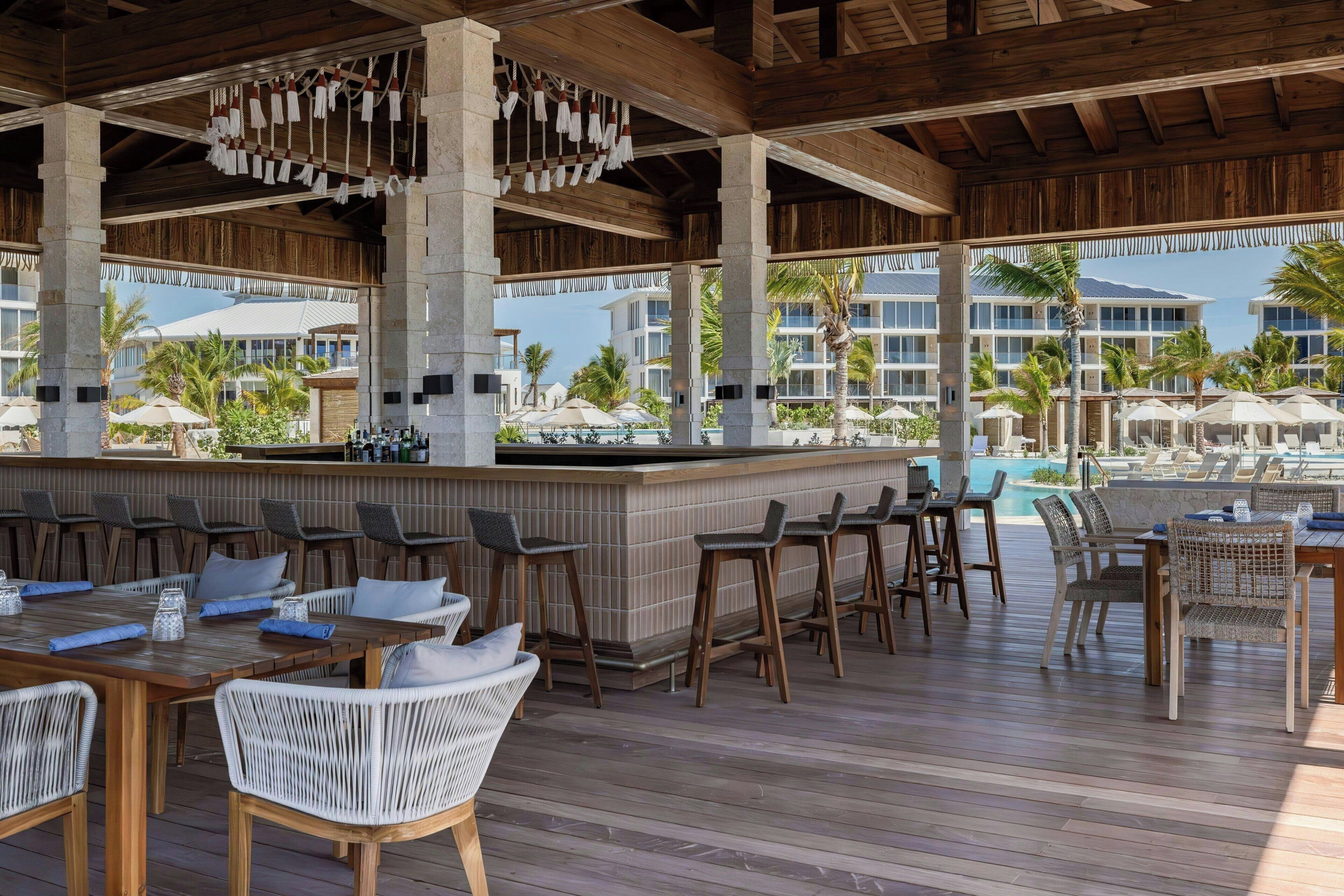 A covered outdoor bar area with wooden furniture, a pool, and a view of resort buildings.