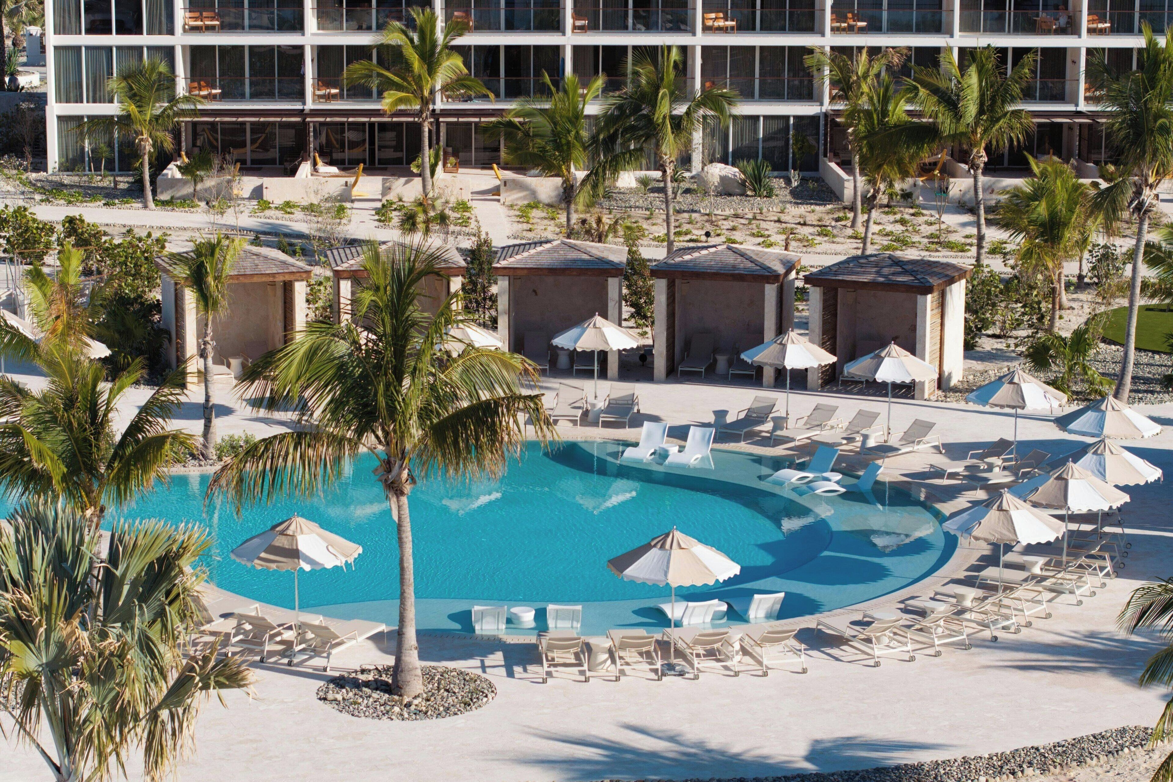 Un resort con una piscina grande rodeada de sillas de descanso y sombrillas, palmeras y un edificio moderno al fondo.