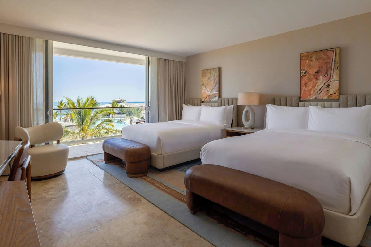 A hotel room with two beds, a desk, a chair, and a view of the ocean.