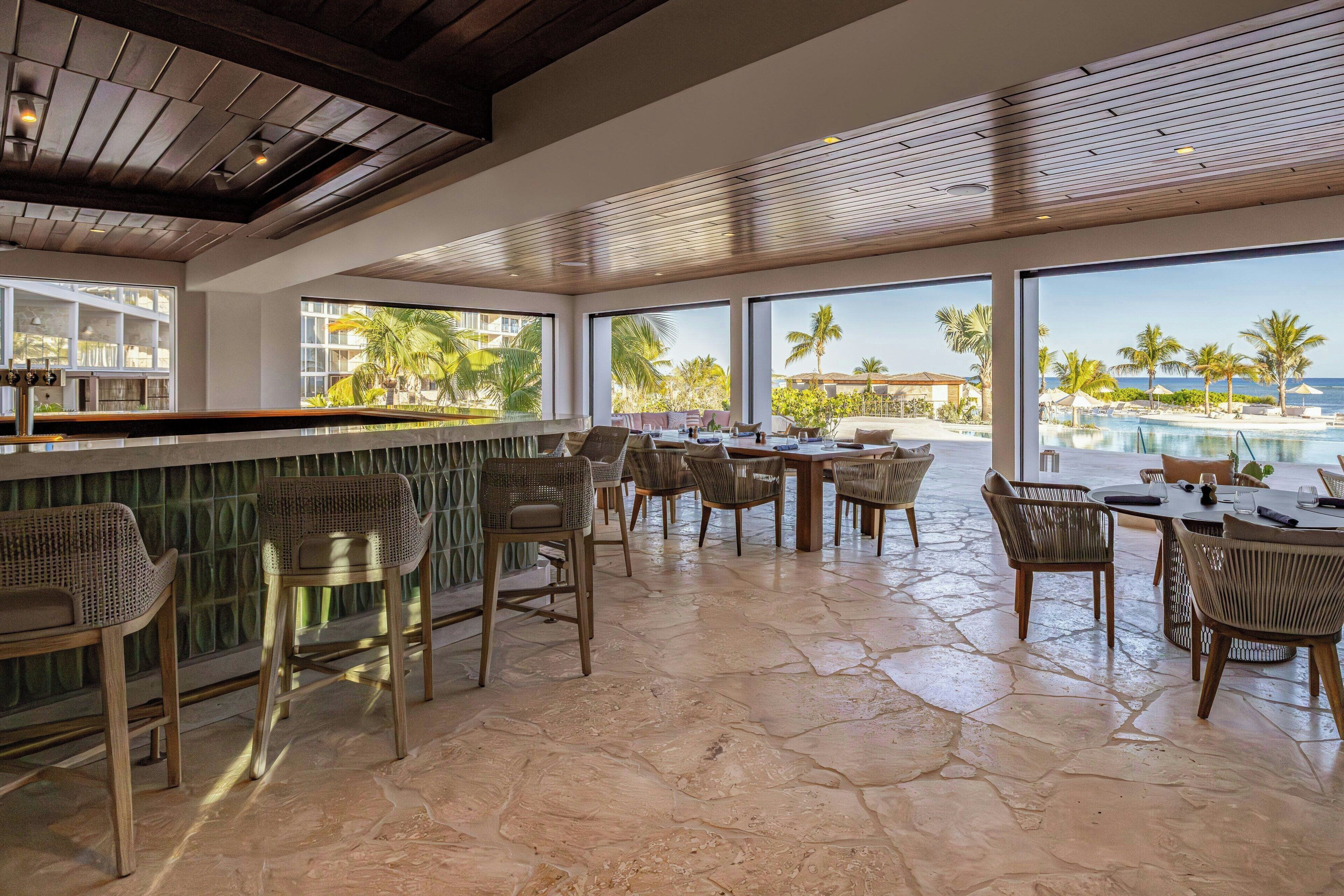 A spacious outdoor seating area with wicker furniture, large windows offering a view of a pool and palm trees, and a wooden ceiling.