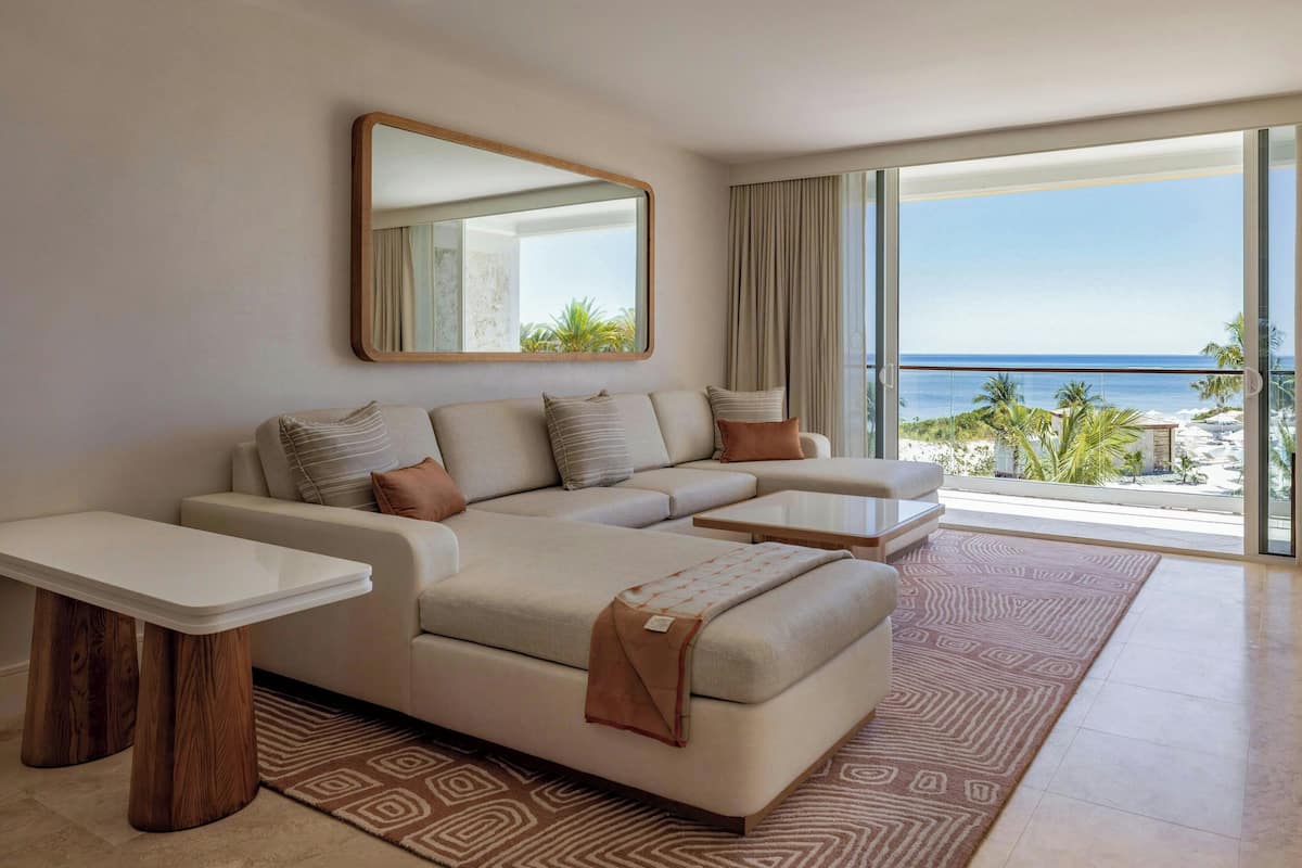 A modern living room with a large sectional sofa, a coffee table, and a side table. A large mirror hangs on the wall, and the room has a view of the ocean.