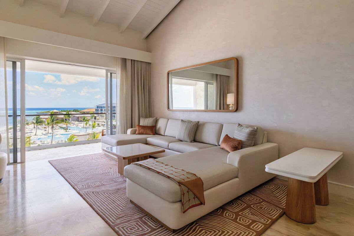 A modern living room with a large sectional sofa, a small coffee table, and a side table. There is a large window offering a view of a pool and palm trees.