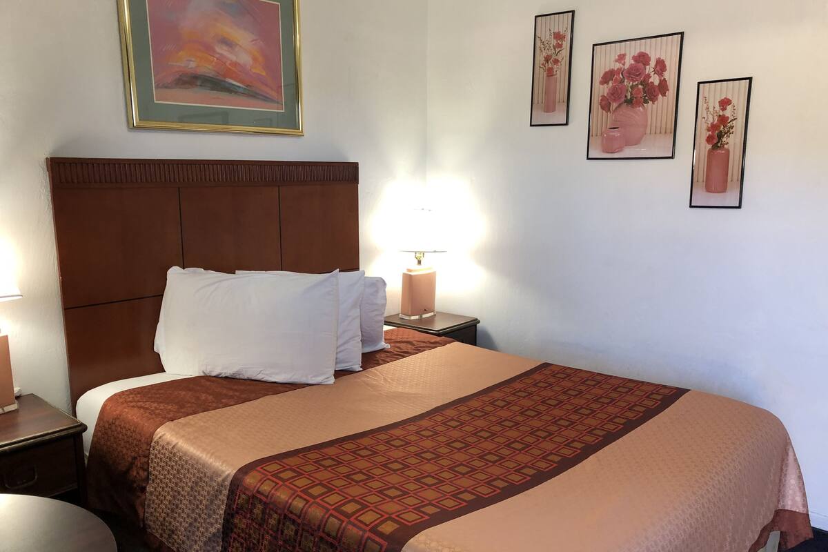 A neatly made bed with a brown headboard, white pillows, and a patterned bedspread. There are two bedside tables with lamps, a round table, and three framed pictures on the wall.