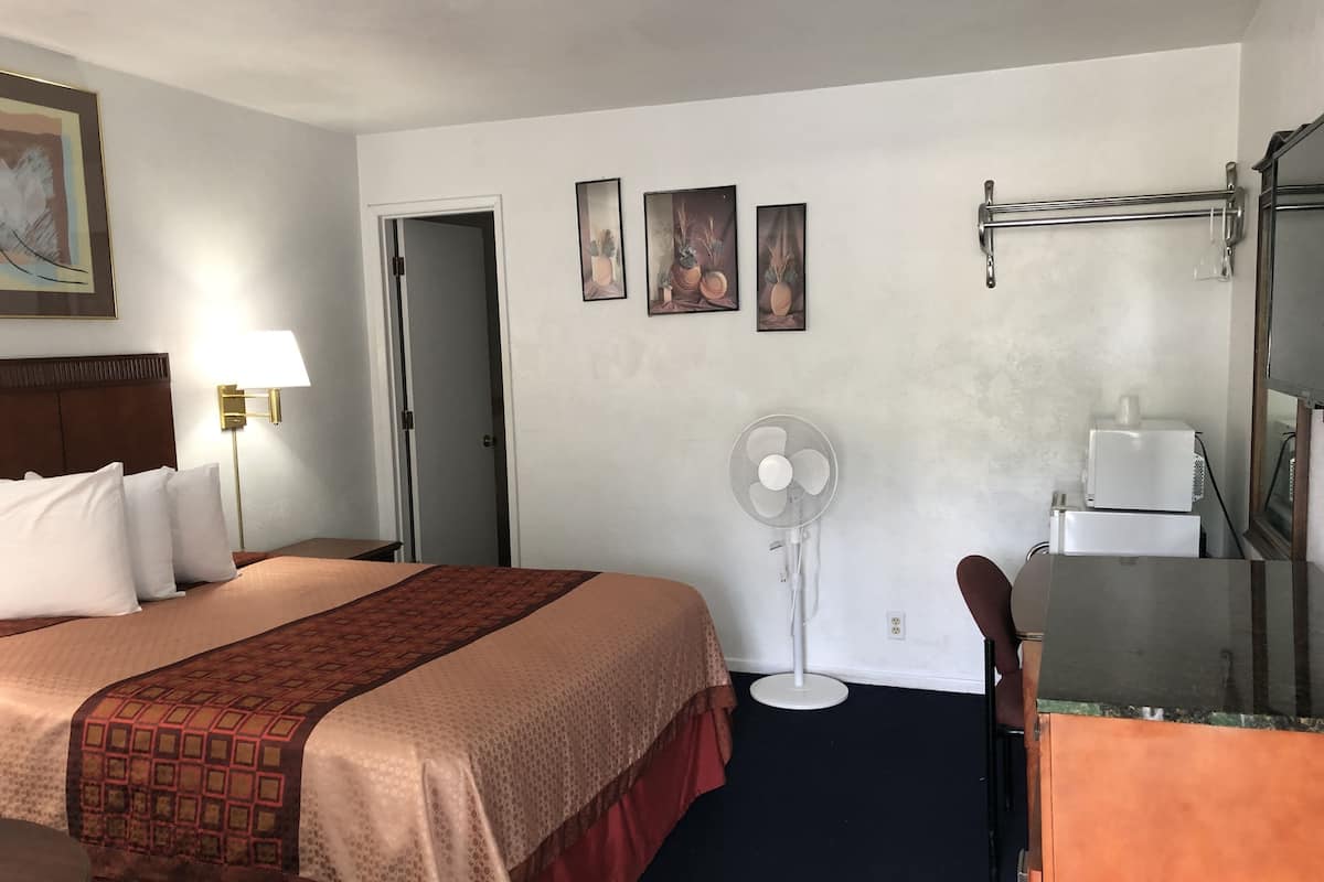 A hotel room with a bed, bedside table, lamp, wall-mounted fan, and a small kitchenette.